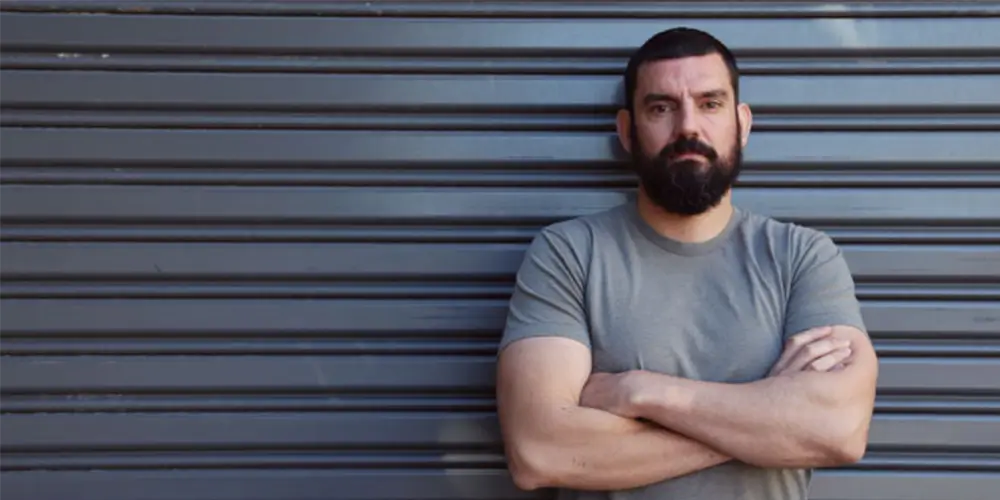 david sutton in grey shirt leaning against wall with arms crossed