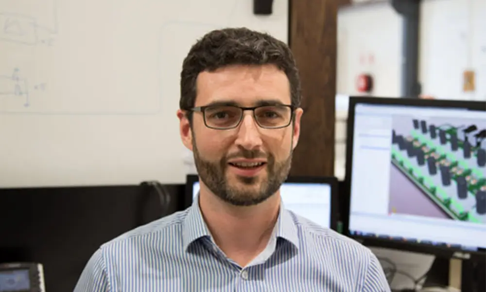 man stands in front of computer screens in button up shirt