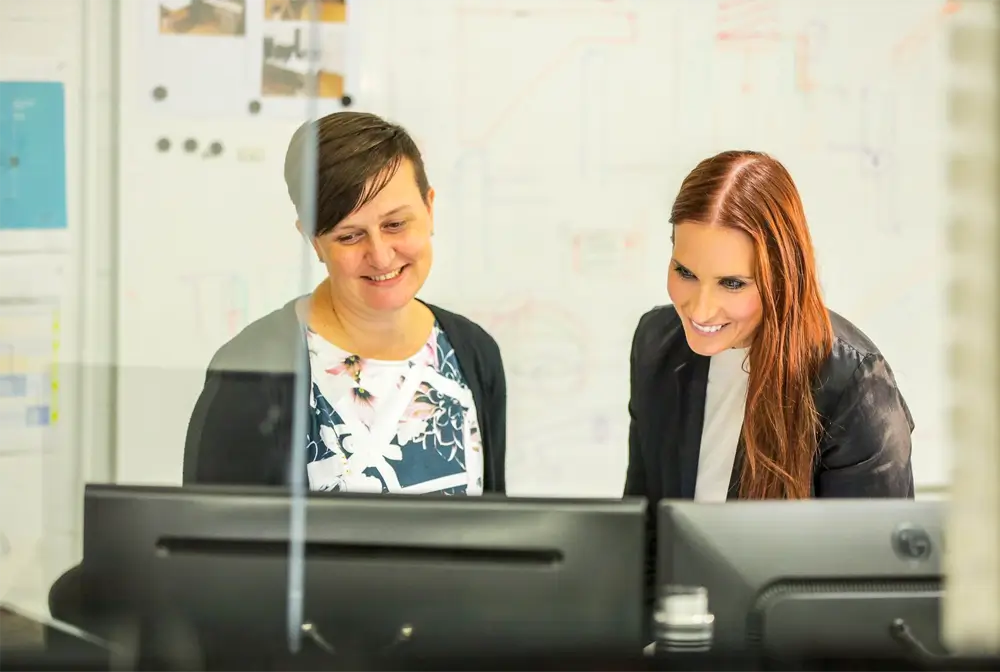 2 women collaborate on their desktop monitors in office